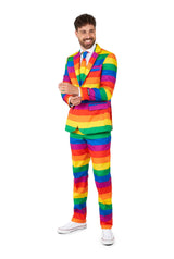 A smiling person wearing the Suitmeister Rainbow suit stands against a white background. The ensemble includes a colorful rainbow-striped jacket and matching pants, paired with white sneakers. They are adjusting their sleeve with one hand.