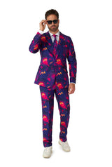 A man wearing the Retro Neon Navy - Carnival suit from Suitmeister, featuring vibrant, abstract patterns, stands against a white background. He is adjusting his sunglasses and sporting a white shirt with a lavender tie, completing the look with white sneakers.