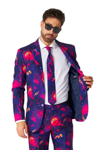 A man dressed in a Retro Neon Navy - Carnival suit from Suitmeister, featuring vibrant cosmic and flamingo patterns. He's accessorized with sunglasses and is holding open one side of his jacket, revealing a white shirt and matching tie underneath.