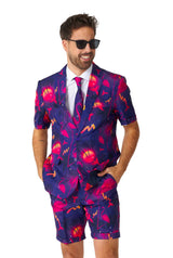 A man wearing sunglasses and the Retro Neon Navy - Carnival suit by Suitmeister, featuring a vibrant cosmic pattern in pink, purple, and red. The suit includes a blazer and matching shorts. He stands with hands in pockets, smiling against a white background.