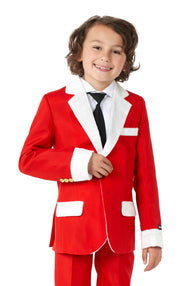 A young child with curly hair is smiling while wearing a Suitmeister Santa Faux Fur Christmas suit, featuring a red jacket with white fur trim, matching pants, and a black tie over a white shirt for a festive holiday look.