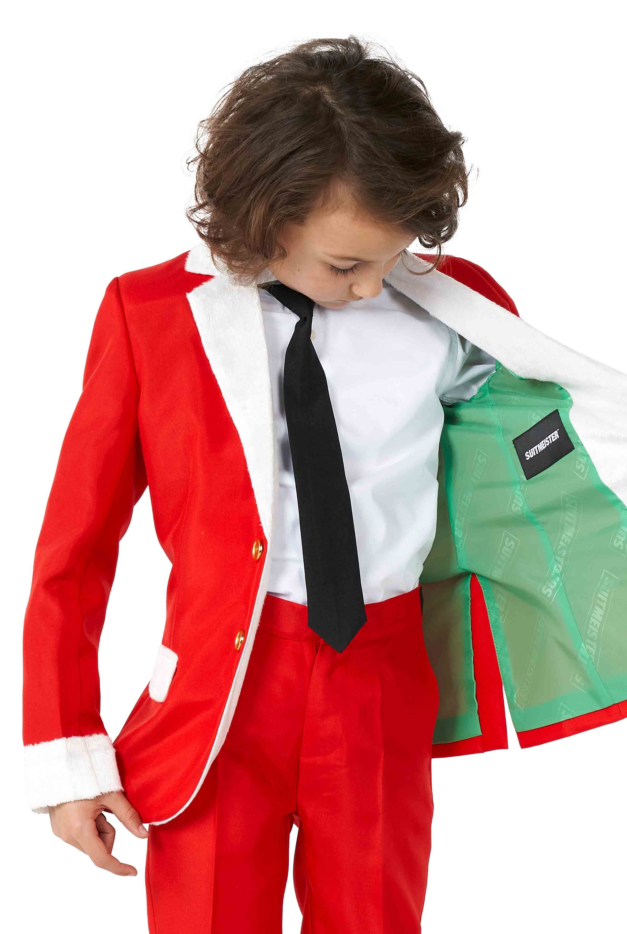 A child wearing the Suitmeister Santa Faux Fur Christmas suit, complete with a white shirt and black tie, peeks inside the jacket to reveal its green lining. The suit is bright red adorned with white faux fur trim on the cuffs and collar.