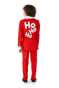 A child, with dark hair, is standing with their back to the camera, wearing a Santa Faux Fur - Christmas suit by Suitmeister. The suit is red and features "Ho Ho Ho" in white letters on the back. They are also wearing black sneakers with white soles.