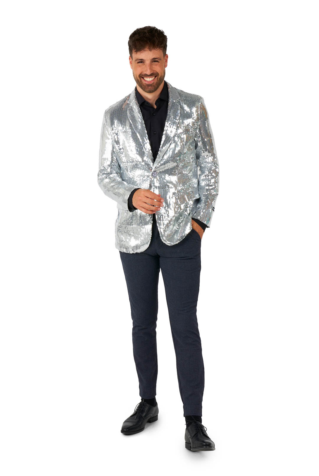 A smiling man stands confidently in a Suitmeister Sequins Blazer Silver, paired with a dark shirt and pants. With one hand in his pocket and holding a small item with the other, he completes the look with black shoes against a white background.