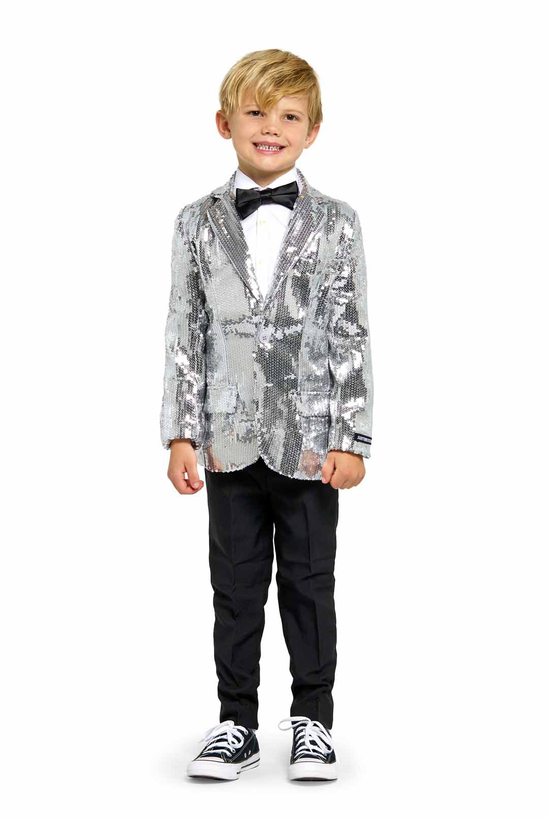 A young boy stands smiling against a plain white background, dressed in Suitmeister's Sequins Silver - Sequin Blazer, complemented by a black bow tie, white shirt, black pants, and black sneakers.