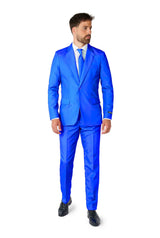 A man in a Solid Blue Suit from Suitmeister stands against a white background, wearing a matching tie and black shoes, and is looking to the side.