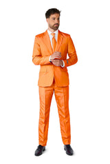 A man wearing a Suitmeister Solid Orange suit, complete with a matching tie and white shirt, stands against a white background. He is adjusting his cuff with one hand, showcasing his trimmed beard and short hair, while wearing black shoes.