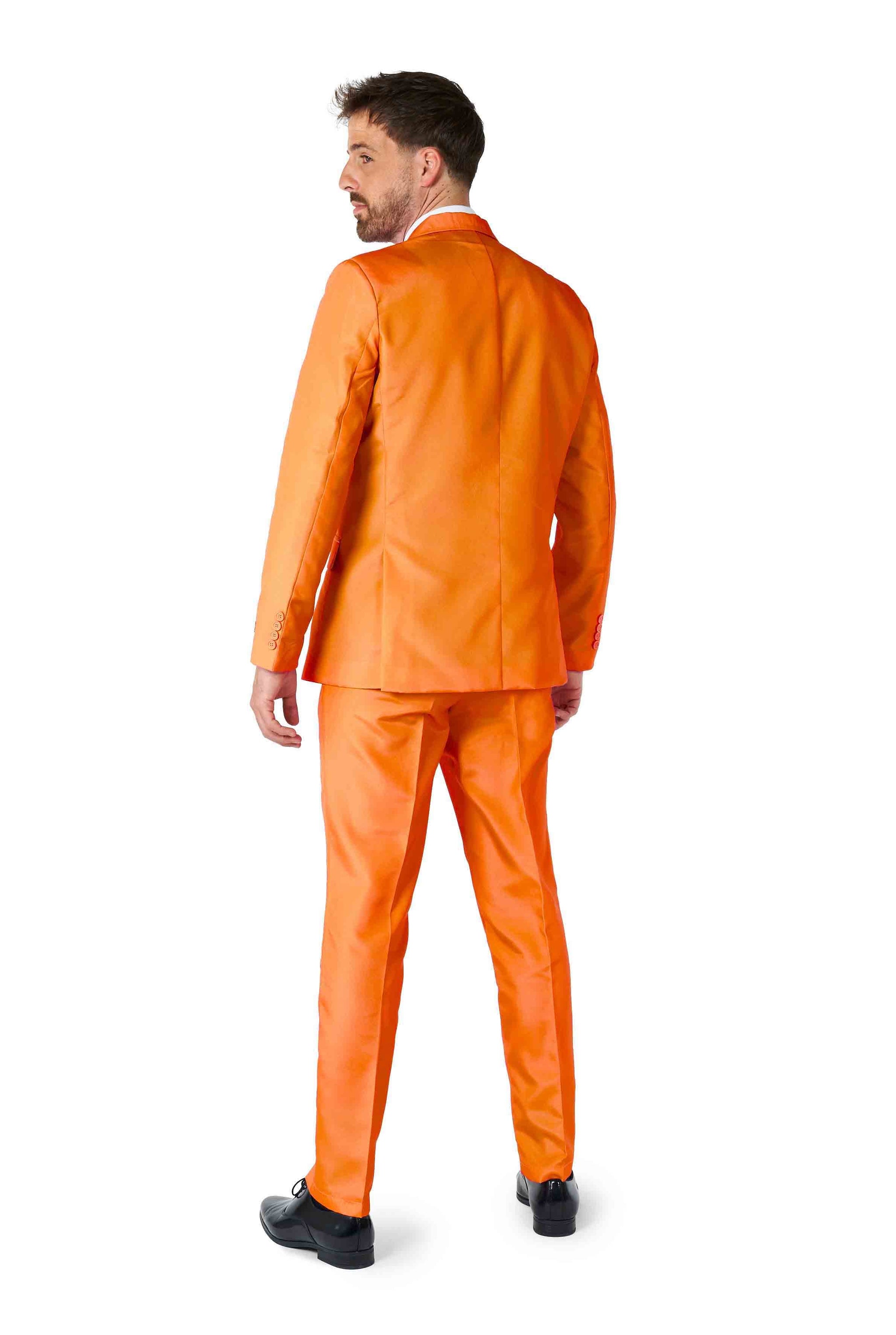 A man wearing the Solid Orange Suit from Suitmeister stands with his back to the camera, slightly turning his head to the side. His black shoes complement the attire, set against a plain white background.