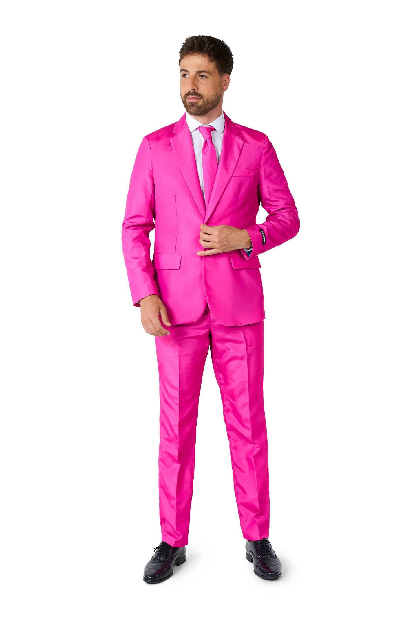 Dressed in Suitmeister's Solid Pink suit, a man with brown hair and a beard exudes confidence. The bright pink suit is complemented by a matching tie and a crisp white dress shirt. He adjusts his jacket with one hand, while his feet are elegantly clad in black dress shoes. All this is set against a plain white background.