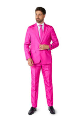 Dressed in Suitmeister's Solid Pink suit, a man with brown hair and a beard exudes confidence. The bright pink suit is complemented by a matching tie and a crisp white dress shirt. He adjusts his jacket with one hand, while his feet are elegantly clad in black dress shoes. All this is set against a plain white background.