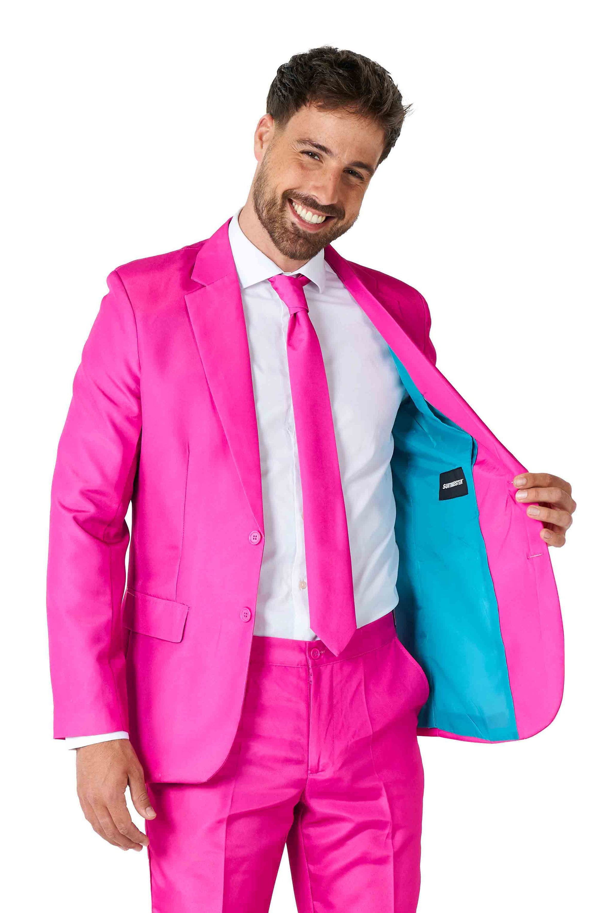 A man wearing the Solid Pink Suit by Suitmeister, complete with a white shirt and pink tie, smiles while holding open his jacket to reveal a light blue lining. He stands in front of a plain white background.