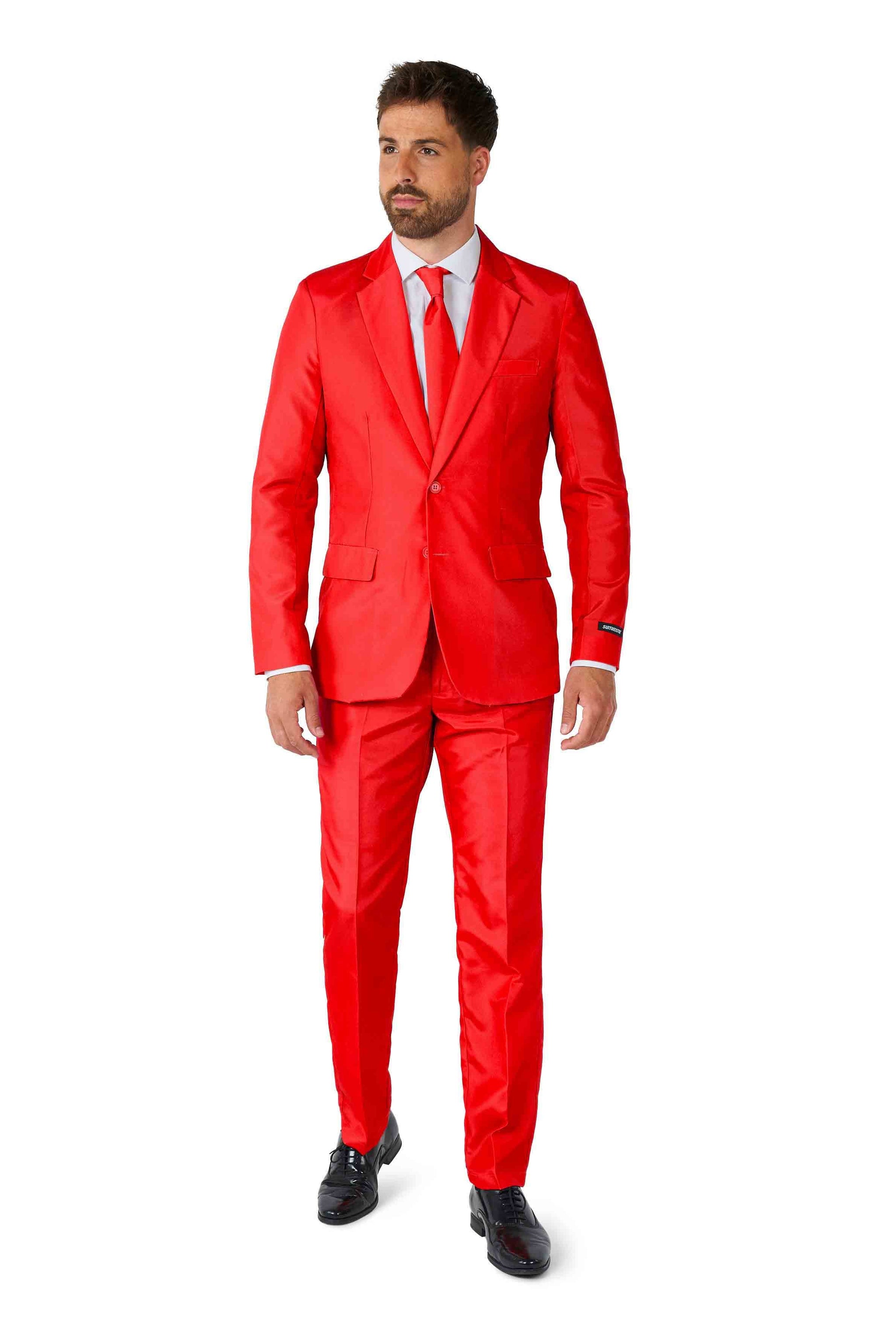 A man is seen dressed in a Suitmeister Solid Red suit, complete with a matching tie and white shirt. Sporting short hair and a beard, he gazes off to the side while wearing black shoes. The backdrop is plain white.