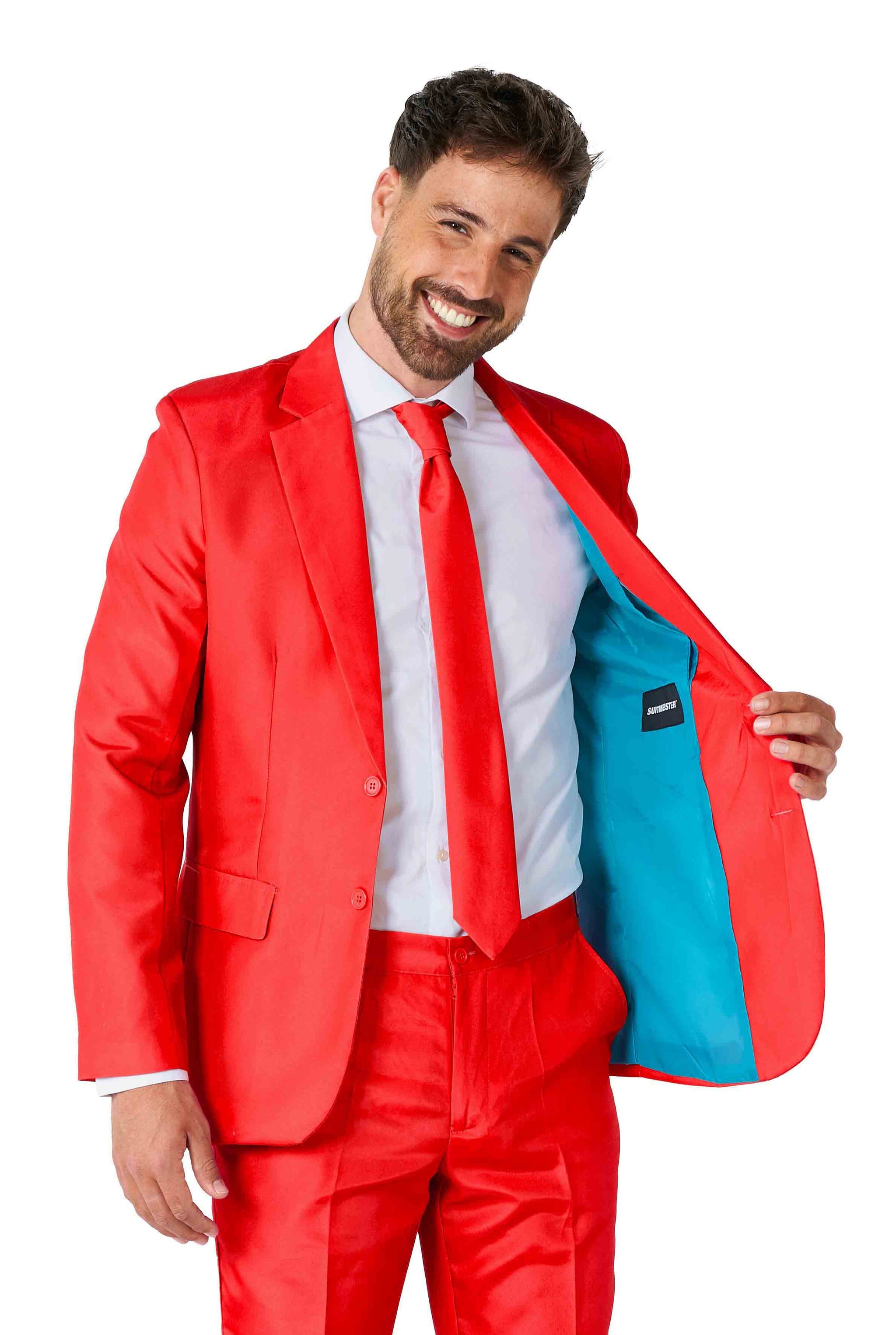 A man wearing a Solid Red suit by Suitmeister, along with a white shirt and red tie, smiles while holding open his jacket to reveal its light blue lining against a plain white background.