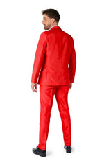 A man in a vibrant Solid Red suit from Suitmeister stands facing away, looking over his shoulder. The suit features a tailored jacket and matching pants. He is wearing black shoes and is set against a plain white background.