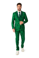 A person dressed in the Suitmeister St. Pats Green suit, featuring a clover pattern, paired with a white shirt and dark green tie, is smiling and walking forward. They are also sporting white sneakers.