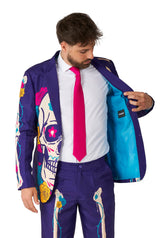 A man is wearing a vibrant Suitmeister's Sugar Skull Purple suit, featuring colorful skull and rose designs on a dark background. He reveals the blue lining of his jacket while sporting a bright pink tie and gazes down at the jacket.