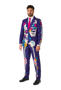 A man stands wearing the Sugar Skull Purple suit by Suitmeister, featuring colorful skeleton patterns and floral designs. He sports a bright pink tie and black shoes.