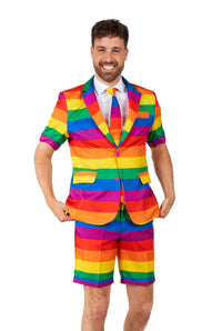 A smiling man wearing the Suitmeister Rainbow suit, which includes a matching tie and shorts, stands against a plain white background. The suit features vibrant horizontal stripes in red, orange, yellow, green, blue, and purple.