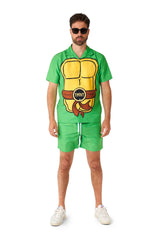 A man sporting sunglasses is dressed in a vivid green TMNT outfit from Suitmeister, adorned with a Teenage Mutant Ninja Turtle motif that includes a yellow shell and a brown belt embossed with TMNT. He pairs the ensemble with white sneakers and stands against a white background.