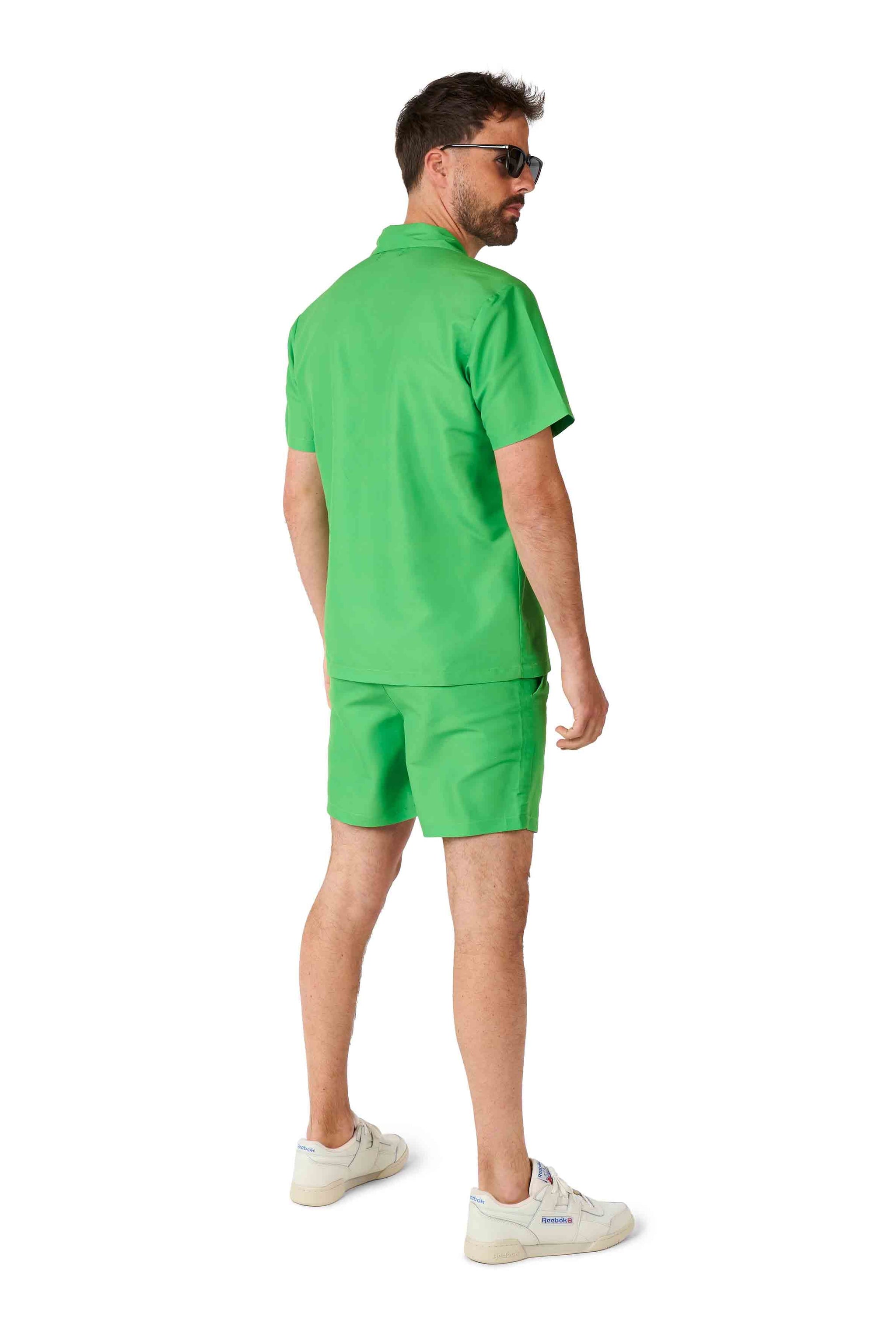 A man wearing sunglasses is standing and looking to the side, dressed in a Suitmeister TMNT outfit, consisting of a matching green short-sleeve shirt and shorts set, paired with white sneakers, against a white background.