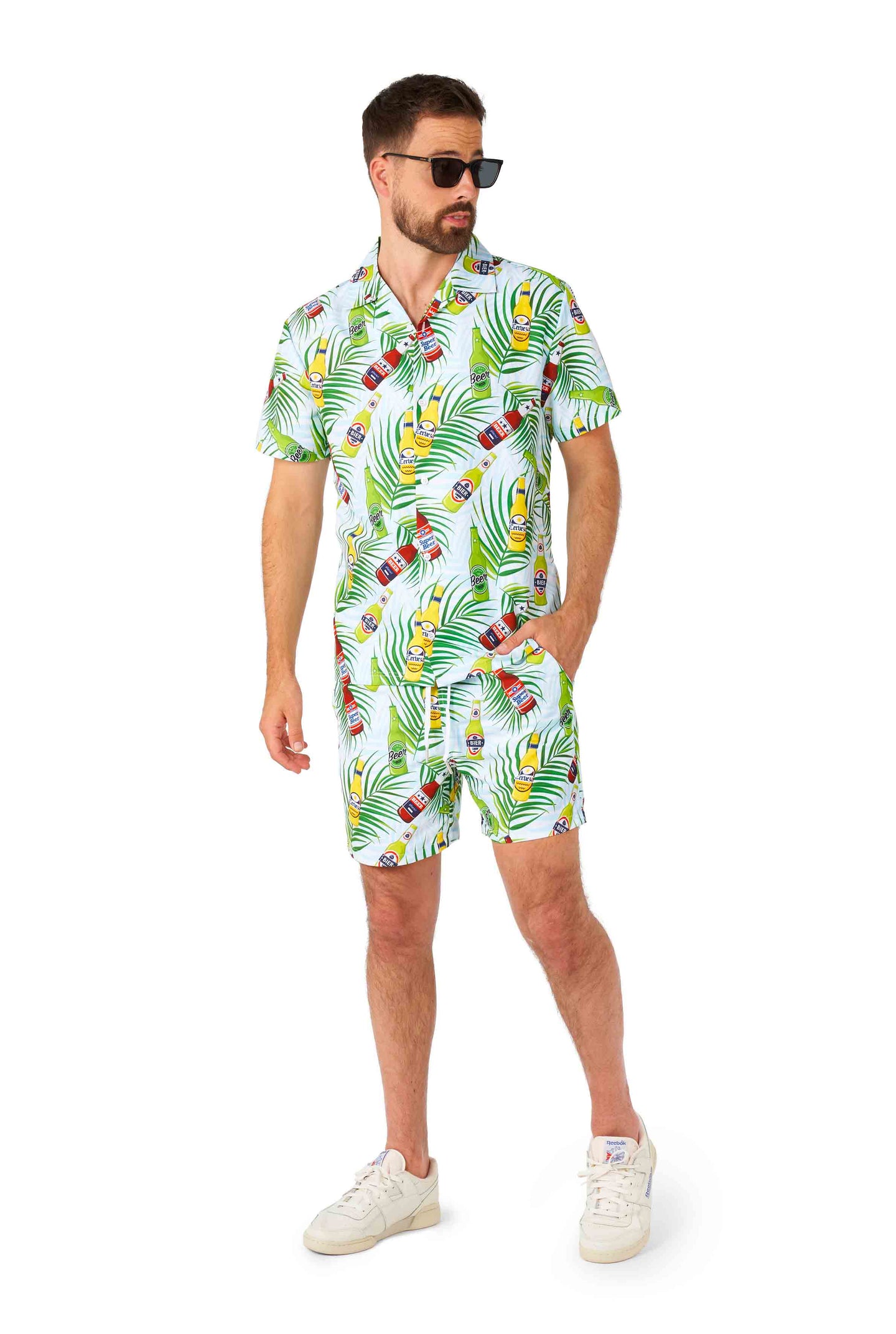 A person wearing sunglasses strikes a pose in a Festival suit named "Tropical Beers Blue" from Suitmeister, showcasing vibrant green leaves and colorful parrots on a matching shirt and shorts set. They stand against a white background with one hand in their pocket.