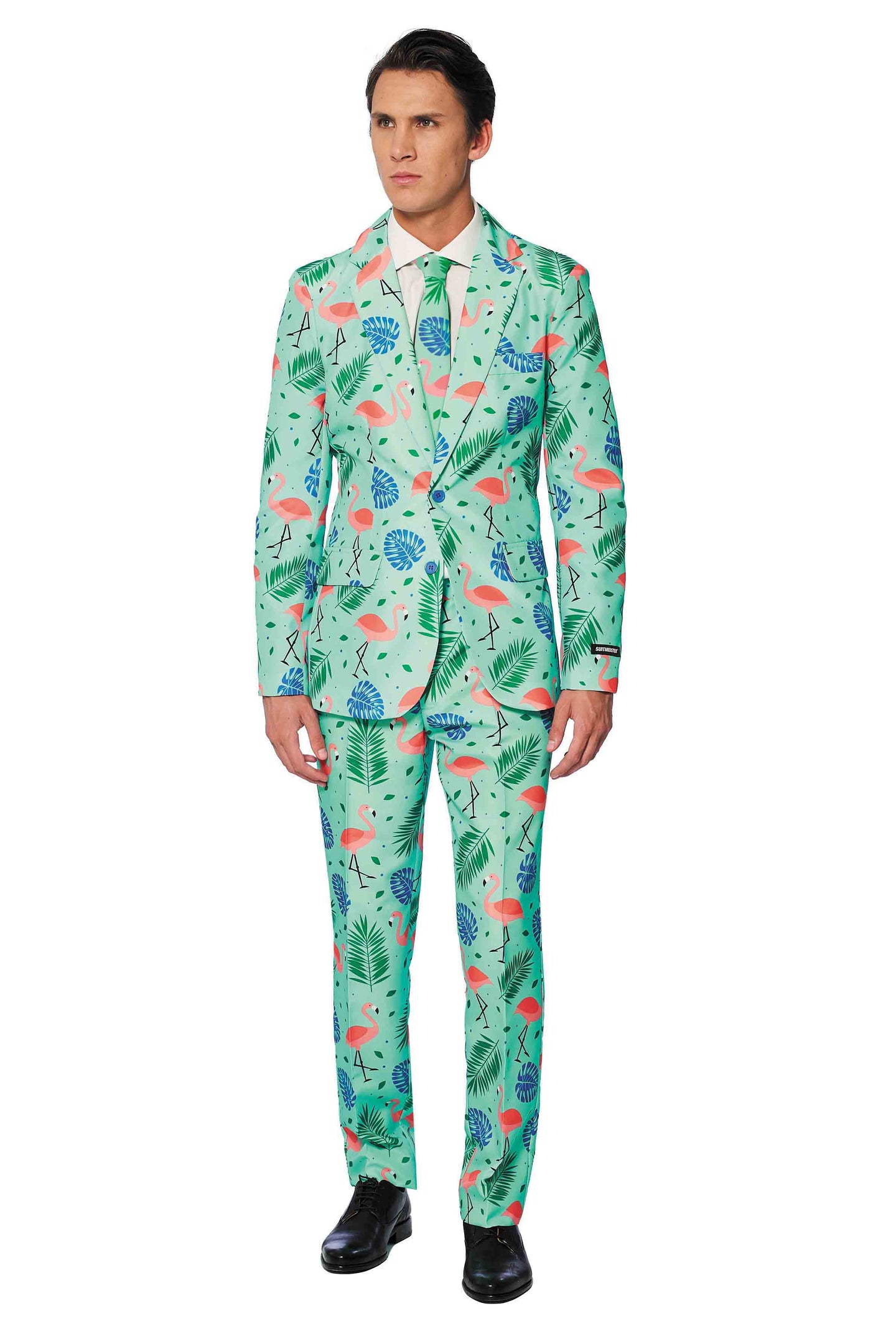 A man stands in a vibrant Suitmeister suit, the Tropical design showcasing pink flamingos and tropical leaves on a light green background. Complementing his look is a white shirt paired with a light green tie and black shoes. The playful pattern contrasts sharply with his serious expression.