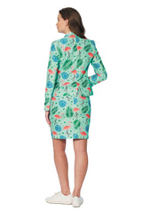 A woman stands facing away, dressed in the Suitmeister's Tropical suit, which includes a green blazer and skirt adorned with a vibrant tropical print featuring flamingos and leaves. She complements this ensemble with white sneakers.