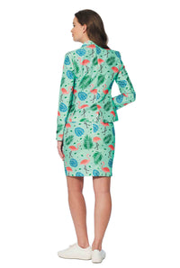 A woman stands facing away, dressed in the Suitmeister's Tropical suit, which includes a green blazer and skirt adorned with a vibrant tropical print featuring flamingos and leaves. She complements this ensemble with white sneakers.