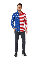 A man with a beard smiles, wearing the USA Flag shirt by Suitmeister, which features an American flag design with stars on the blue side and red and white stripes on the other. With one hand in his pocket, he pairs it with dark pants and black shoes while standing against a white background.