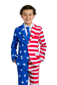 A boy beams with joy as he wears the Suitmeister USA Flag - 4th of July outfit, distinguished by its blue side adorned with white stars and the opposite side showcasing red and white stripes. His hands rest casually in his pockets as he stands against a plain white backdrop.