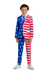 A young boy stands smiling, donning the Suitmeister USA Flag - 4th of July outfit. The suit showcases stars on a blue background on one side and red and white stripes on the other, perfectly paired with his white sneakers and a blue tie.