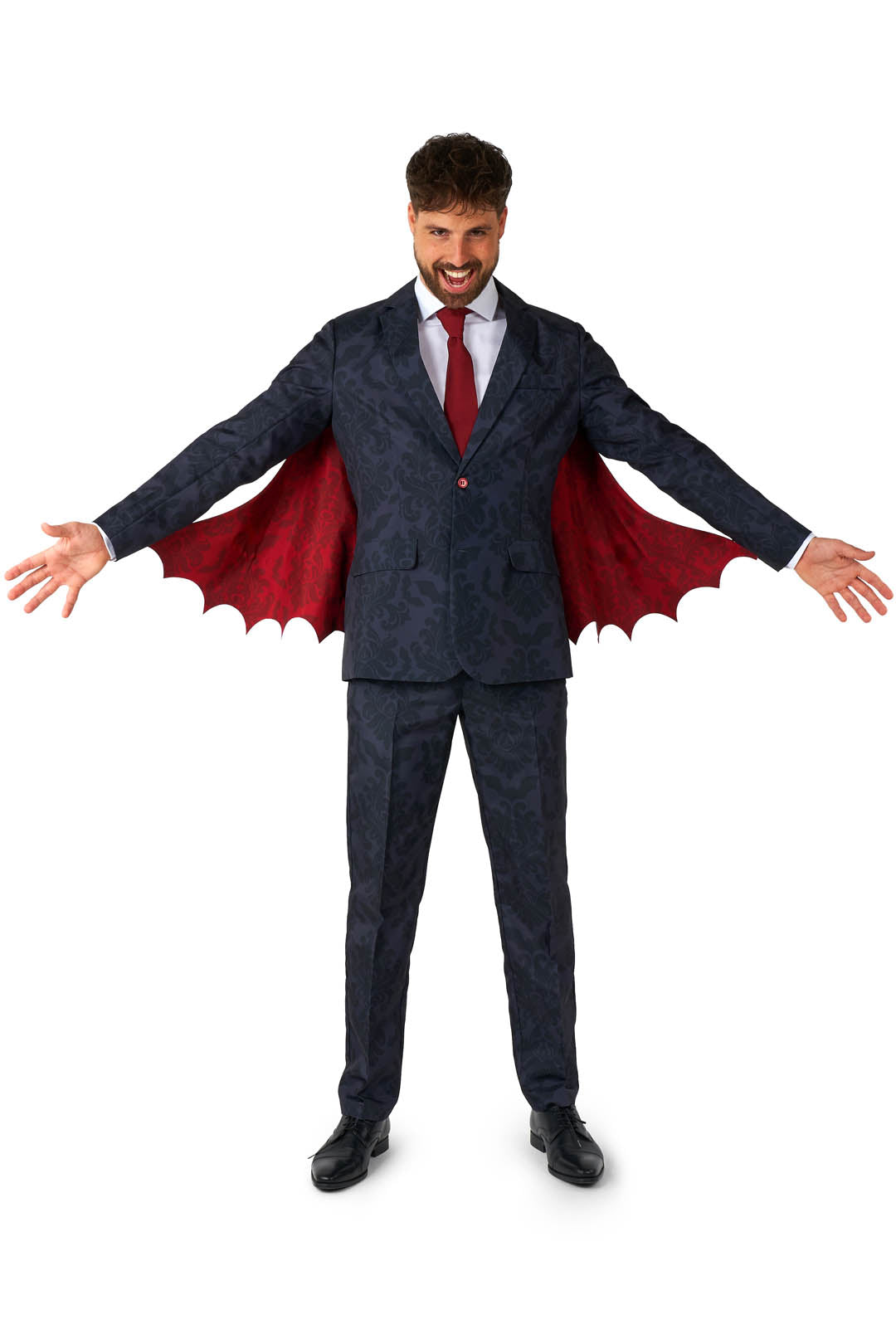 A man in the Victorian Vampire Suit Black from Suitmeister stands with his arms outstretched, showcasing a dark suit with an intricate patterned design and a red tie. The suit jacket features dramatic red, bat-like wing extensions. His playful expression, complete with an open mouth, adds to the theatrical style.